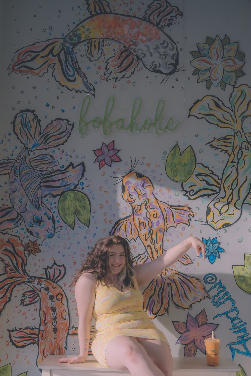 picture of artist madeline in front of a fish mural she painted around a bobaholic sign